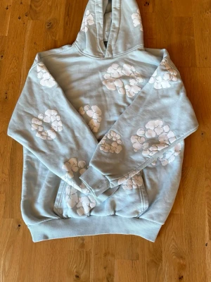 Denim tears hoodie - Säljer en ljusblå hoodie från Denim Tears i storlek XS. Hoodien har vita bomullsliknande tryck över hela plagget, klassisk huva och ribbade muddar vid ärmar och nederkant. Perfekt för dig som gillar streetwear och unika prints.