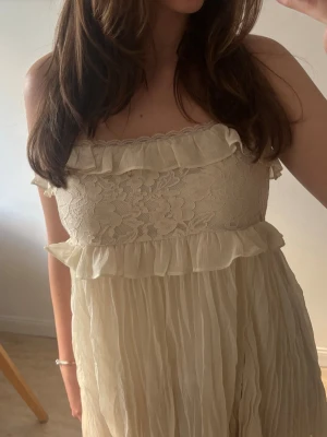 Beige axelbandsklänning med spets - Söt beige klänning med tunna axelband, volangdetaljer och spets upptill. Överdelen har ett blommigt spetsmönster och volanger vid bysten, medan kjolen är plisserad och luftig. Perfekt för sommarens varma dagar.