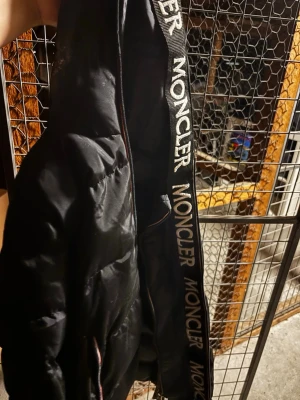 Svart dunväst från Moncler - Säljer en svart dunväst från Moncler med breda kanaler och tydlig logga på insidan av dragkedjan. Västen har en sportig look med Moncler-tryck längs kanten och är tillverkad i glansigt material. Perfekt för dig som vill ha något stilrent och trendigt.