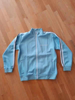 Ljusblå track jacket från Palm Angels - Snygg ljusblå track jacket från Palm Angels med vita och svarta ränder längs ärmarna. Jackan har dragkedja framtill, ståkrage och två fickor med dragkedja. Materialet är troligtvis polyester, perfekt för sportiga outfits. Den är i storlek M men passar även L