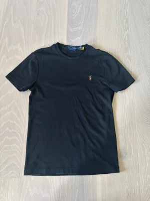 Svart t-shirt från Polo Ralph Lauren - Klassisk svart t-shirt från Polo Ralph Lauren med rund hals och den ikoniska broderade polospelaren på bröstet. Tillverkad i mjuk bomull och har en normal passform som funkar till det mesta. Enkel och stilren design som alltid är rätt. Vid intresse är det bara att höra av sig 