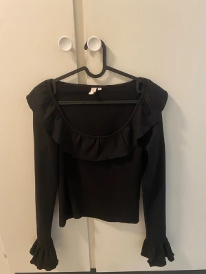 Svart långärmad topp med volang - Snygg svart långärmad topp med bred volang runt ringningen och volangdetaljer vid ärmsluten. Toppen har en offshoulder-design och är tillverkad i ett mjukt, stretchigt material som sitter bekvämt på kroppen.