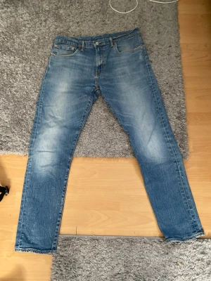 Ljusblå raka jeans med slitningar Levis  - Säljer ett par ljusblå jeans med raka ben och diskreta slitningar på framsidan. Jeansen har klassisk femficksmodell och knappgylf. Materialet är denim i bomull och passformen är avslappnad men ändå smal.