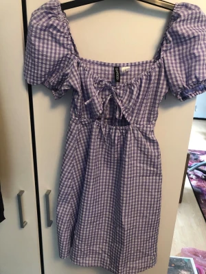 Lila rutig klänning med Dorothy känsla - Superfin lila och vit rutig klänning från H&M Divided med puffärmar, snörning framtill och dragkedja på sidan och är i en söt midilängd. Klänningen har en omlottkänsla upptill och är perfekt för vår och sommar eller höst med ett par leggings.
