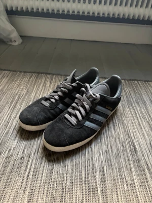Adidas Gazelle svarta sneakers - Klassiska Adidas Gazelle sneakers i svart mocka med gråa ränder och snören. Låg modell med vit sula och diskret guldfärgad Gazelle-text på sidan. Perfekta för en stilren och sportig look. Hör av dig vid frågor// kan gå ner i pris vid snabb affär!😃/ Milton