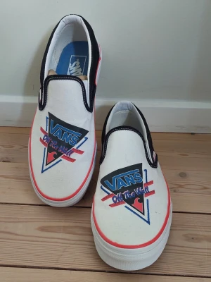 Vans slip-on sneakers med retrotryck - Vita slip-on sneakers från Vans med svart och röd detalj, samt retroinspirerat tryck på ovandelen med texten 'Off the Wall'. Skorna har svart kant och vit sula. Tillverkade i canvasmaterial och har klassisk Vans-logga på sidan. Oanvända men ett litet märke, se bild.