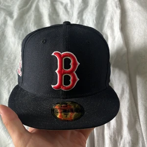 Svart/mörkblå Boston Red Sox keps från New Era - Säljer en svart/mörkblå 59FIFTY keps från New Era med Boston Red Sox logga broderad i rött och vitt framtill. Kepsen har platt skärm, broderad MLB-logga bak och texten 'Est. 2009' på sidan. Klassisk modell i bomull, perfekt för dig som gillar sportig stil. Storlek 59.6 cm