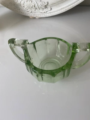 Grön sockerskål i pressglas - Säljer en vintage grön sockerskål i pressglas med två handtag och dekorativt stjärnmönster i botten. Skålen har en åttakantig form och genomskinlig grön färg som ger en retro känsla till dukningen.vet ej om det är uran eller mangan 
