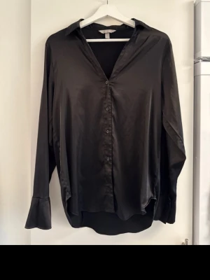 Svart skjorta/blus i siden - Stilren svart skjorta i silkesliknande material med klassisk krage och knappar framtill. Skjortan har lång ärm med bred manschett och en loose passform som ger en avslappnad men ändå snygg look. Perfekt att styla med jeans eller kjol.