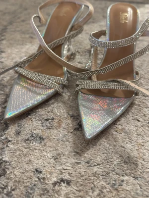 Silvriga holografiska pumps med strass - Säljer ett par silvriga pumps med holografisk yta och spetsig tå. Skorna har flera tunna remmar dekorerade med strass och en transparent klack som ger en futuristisk vibe. Perfekta för dig som vill sticka ut och glänsa på nästa event.