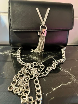Valentino handväska med silverdetaljer festväska - Valentin svart handväska i slätt material med en stor silverfärgad kedja och dekorativt V-spänne med hängande kedjor framtill. Väskan har ett innerfack och stängs med lock. Perfekt accessoar för att lyfta din outfit.