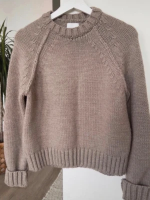 Beige stickad tröja från H&M - Mysig beige stickad tröja från H&M med rund halsringning och ribbade muddar. Tröjan har diskreta flätade detaljer längs axlarna och är tillverkad i ett tjockare garn som ger en avslappnad och trendig look.