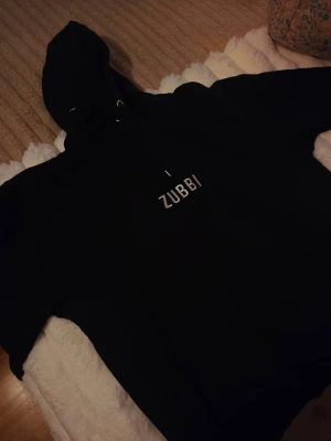 Merch ODZ hoodie  Dunderkläder - Säljer en svart hoodie från Dunderkläder med texten 'ZUBBI' tryckt på bröstet. Tröjan har en luva och långa ärmar.   Storlek L / XL
