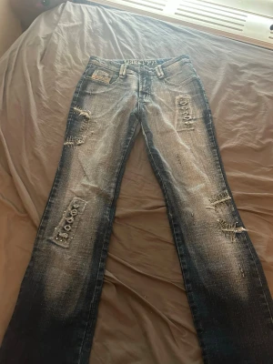 Blå slitna jeans med patchdetaljer - Säljer ett par blå jeans med slitningar och patchdetaljer framtill. Jeansen har raka ben, ljusa tvättade partier och coola snörningsdetaljer på bakfickorna. Materialet är klassisk denim och passformen är relaxed. Perfekta för en avslappnad streetstyle-look. Vet inte märket på dessa men dem sitter bra på.