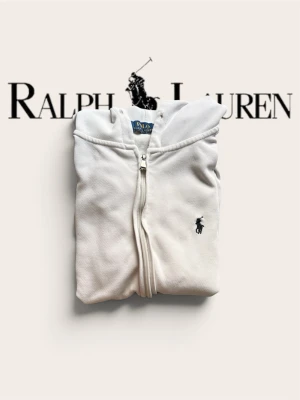 Vit hoodie från Polo Ralph Lauren - Säljer en stilren vit hoodie från Polo Ralph Lauren med dragkedja framtill och klassisk huva. Hoodien har det ikoniska broderade logotypen på bröstet, ribbade muddar och fickor på sidorna. Perfekt för en clean och avslappnad look.(Kommer med snöre)