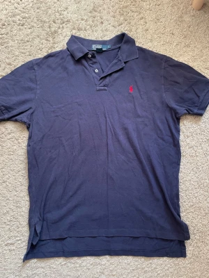 Mörkblå pikétröja från Polo Ralph Lauren - Klassisk mörkblå pikétröja från Polo Ralph Lauren med röd broderad logga på bröstet. Tröjan har krage, knappar vid halsen och korta ärmar. Tillverkad i mjuk bomull. Kolla noga på alla bilder!❗️