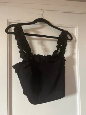 Svart linnetopp med volangdetaljer - Snygg svart linnetopp med breda axelband och volangdetaljer längs ringningen och axelbanden. Toppen är ärmlös och har en croppad passform som ger en trendig look. Perfekt att styla med höga jeans eller kjol.