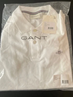 Vit pikétröja från GANT - Klassisk vit pikétröja från GANT med broderad logga på bröstet och knappslå framtill. Tröjan har krage och korta ärmar, perfekt för en clean och stilren look. Tillverkad i mjuk bomull för skön komfort.