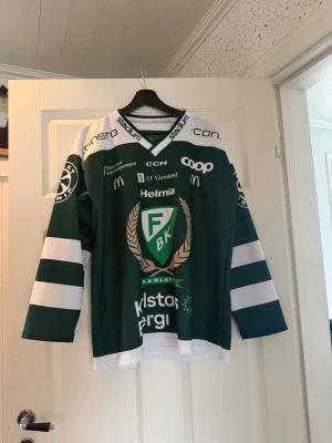Färjestad BK hockeytröja grön/vit - Säljer en grön och vit hockeytröja från Färjestad BK med klubbmärke och flera sponsorer tryckta på både fram- och baksida. Tröjan har långa ärmar med vita ränder och V-ringad krage. Perfekt för dig som vill stötta laget på match eller träning.