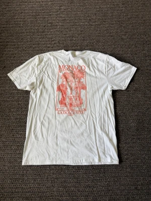 Vit t-shirt med Monaco-tryck - Säljer en vit t-shirt med rött tryck på ryggen som föreställer palmer och en byggnad samt texten 'MONACO LA DOLCE VITA RESORT'. T-shirten har klassisk passform och korta ärmar. Perfekt för dig som gillar grafiska motiv och semesterkänsla.