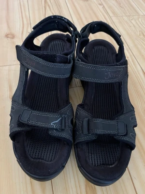 Svarta sandaler med kardborre från Joya - Svarta sandaler från Joya med sportig design, justerbara kardborreband och vadderad innersula. Tillverkade i syntet och textil med kontrastsömmar och stabil sula för extra komfort. Perfekta för dig som gillar funktionella och stilrena skor.
