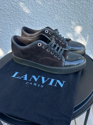 Bruna Lanvin sneakers i mocka och lack - Typ nya as bra skick kan passa 40-41 Snygga bruna sneakers från Lanvin med ovandel i mocka och tåparti i svart lack. Skorna har svarta skosnören, metallfärgade öljetter och en mörk sula. Perfekta för dig som gillar stilrena och exklusiva detaljer.