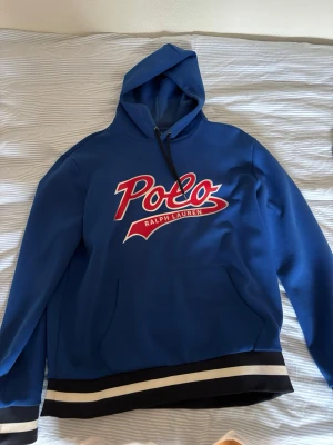 Blå hoodie från Polo Ralph Lauren - Snygg blå hoodie från Polo Ralph Lauren med stor röd och vit logga på bröstet. Huva med svarta snören och känguruficka framtill. Ribbstickade muddar och svart/vita ränder längst ner. Perfekt för en sportig och avslappnad stil.