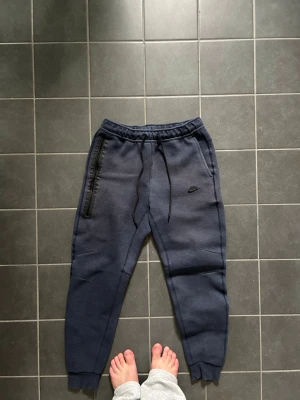 Mörkblå Nike mjukisbyxor - Säljer ett par mörkblå mjukisbyxor från Nike med smal passform och resår i midjan. Byxorna har dragsko, ribbade muddar vid bensluten och en svart dragkedjeficka på sidan. Perfekta för chill eller träning.