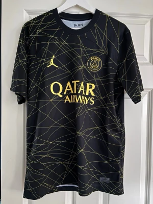 PSG Mbappé matchtröja svart/gul - Snygg PSG matchtröja i svart med gula detaljer och grafiskt mönster. Framsidan har Qatar Airways-logga och Paris Saint-Germain-emblem, baksidan har Mbappé och nummer 7. Tillverkad i lätt och ventilerande material, perfekt för fotboll eller träning.