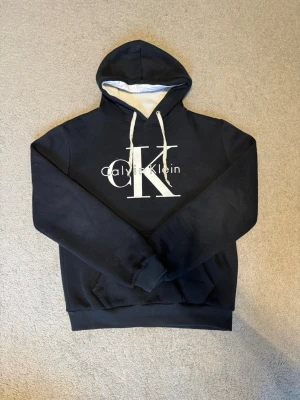 Dark blue hoodie Calvin Klein - Snygg svart hoodie från Calvin Klein med stor vit CK-logga och text på bröstet. Hoodien har vit dragsko i huvan och mjukt foder på insidan av huvan. Klassisk passform med ribbade muddar vid ärmslut och nederkant.