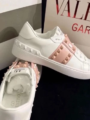 Valentino Rockstud vita sneakers - Snygga vita sneakers från Valentino med ljusrosa band och ikoniska nitar i matchande färg. Skorna har en låg profil och är tillverkade i läder med detaljerade sömmar och robust sula. Perfekta för dig som gillar en edgy men stilren look.