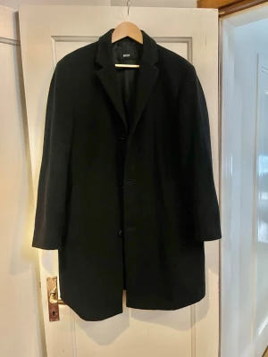 Hugo boss ullrock - Stilren svart ullkappa med rak passform och enkelknäppning. Kappan har klassisk krage och dolda sidofickor, perfekt för en clean och tidlös look. Passar till lager på lager och funkar lika bra till jeans som till kostymbyxor. Storlek M