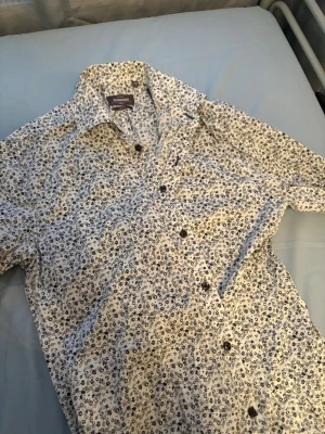 Blommig skjorta slim fit  - Säljer en vit skjorta från Dressmann med svart blommigt mönster. Skjortan har slim fit-passform, långa ärmar och knappar framtill. Perfekt för dig som gillar stilrena detaljer och stretchigt material.