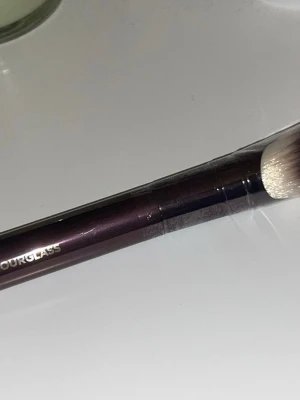 Hourglass vinklad sminkborste - Snygg vinklad sminkborste från Hourglass med mörklila metallhandtag och täta, mjuka syntetiska borst i vitt och grått. Perfekt för contouring eller foundation. Handtaget har en elegant metallfinish och borsten känns lyxig och proffsig.