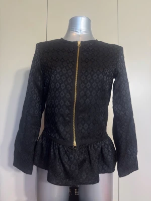 Svart mönstrad jacka med volang - Snygg svart jacka med diskret diamantmönster och guldfärgad dragkedja framtill. Jackan har rund halsringning, långa ärmar och en volang nedtill som ger en feminin touch. Materialet är strukturerat och känns lite tjockare, perfekt för lager på lager.