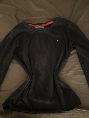 Svart långärmad tröja Tommy Hilfiger - Klassisk marinblå långärmad tröja från Tommy Hilfiger med rund halsringning och liten broderad logga på bröstet. Tröjan har en enkel och stilren design med röd detalj på insidan av kragen. Perfekt till jeans eller kjol.