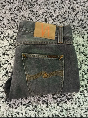 Grå jeans från Nudie Jeans - Säljer ett par grå jeans från Nudie Jeans med klassisk femficksmodell och gul kontrastsöm på bakfickorna. Jeansen har rak passform och är tillverkade i denim med en snygg tvättad look. Perfekta för dig som gillar stilrena och tidlösa jeans.