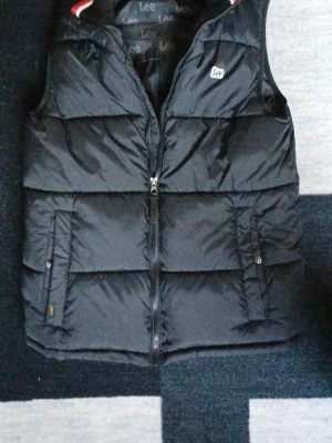 Svart dunväst från Lee - Snygg svart dunväst från Lee med quiltad design och dragkedja framtill. Västen har två fickor med knappstängning. Inga defekter är bara använd ca 10 gånger. Skriv om ni har frågor!