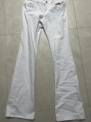 Vita bootcut jeans med fickdetalj - Snygga vita bootcut jeans med klassisk femficksdesign och en extra ficka med lock bak. Jeansen är låg midjas  och vida ben, perfekta för en trendig look. Tillverkade i ett mjukt denimtyg som ger en bekväm känsla hela dagen.