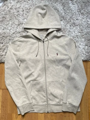 Ljusgrå hoodie från Polo Ralph Lauren - Ljusgrå hoodie med dragkedja från Polo Ralph Lauren. Klassisk design med huva och dragsnören, två fickor framtill och den ikoniska broderade loggan på bröstet. Tillverkad i mjuk bomullsblandning för maximal komfort.