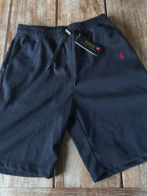 Mörk blåa från Polo Ralph Lauren - Svarta shorts från Polo Ralph Lauren med elastisk midja och snörning. Klassisk röd broderad logga på ena benet och mjukt material som känns skönt mot huden. Perfekta för sommaren och avslappnade dagar.
