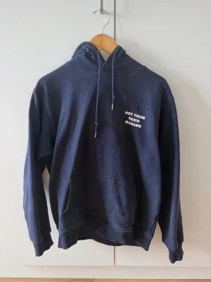 Drole de Monsieur NOT FROM PARIS MADAME hoodie - Säljer en mörkblå hoodie från Drole De Monsieur med vit text 'NOT FROM PARIS MADAME' på bröstet och ryggen. Hoodien har huva med dragsko och en stor magficka. Tillverkad i mjukt bomullsmaterial och har ribbade muddar vid ärmar och nederkant. Perfekt för en avslappnad och trendig look.