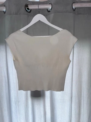Beige ribbad crop top med v-ringning - Säljer en beige ribbad crop top med v-ringning framtill och breda axelband. Toppen är stretchig och har en rak nederkant med lätt vågig kant. Perfekt att styla till jeans eller kjol för en enkel och trendig look.