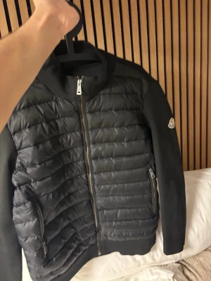 Svart pufferjacka från Moncler - Snygg svart pufferjacka från Moncler med quiltad front och släta ärmar. Jackan har dragkedja framtill, två sidofickor med dragkedja och en klassisk Moncler-logga på ärmen. Insidan har ett unikt serietidningsmönster. Perfekt för kalla vinterdagar.