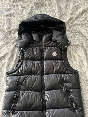Svart dunväst med huva från Moncler - Snygg svart dunväst från Moncler med glansig finish och avtagbar huva. Västen har två dragkedjeförsedda fickor fram och Moncler-logga på bröstet. Perfekt för lager-på-lager och håller dig varm under kyliga dagar.