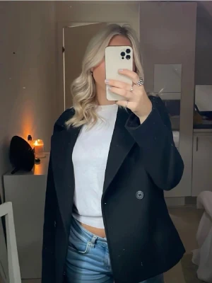 Svart kappa med bred krage - Stilren svart kappa med breda slag och enkelknäppning. Modellen är rak och något kortare, vilket ger en modern look. Perfekt att slänga över en hoodie eller stickad tröja. Kappan har en klassisk design och passar till många olika outfits.