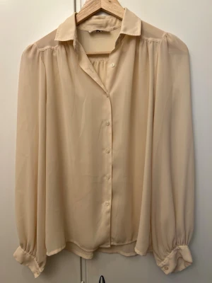Beige blus från Daniel Duval Paris - Stilren beige blus från Daniel Duval Paris med klassisk krage och knäppning framtill. Blusen har långa, puffiga ärmar med breda manschetter och knappar. Tillverkad i ett tunt, lätt material som ger ett elegant fall.
