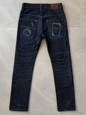Nudie Jeans 29/30 - Säljer ett par mörkblå jeans från Nudie Jeans med skön passform och klassiska fem fickor. Jeansen har coola slitningar och unika sömmar på bakfickorna. Tillverkade i denim med en snygg tvättad look. Perfekta för dig som gillar stilrena men ändå personliga jeans.