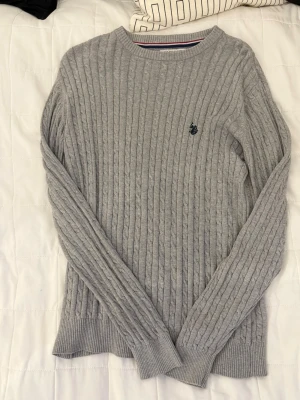 Grå kabelstickad tröja polo assn  - Snygg grå kabelstickad tröja med rund hals och lång ärm. Tröjan har ett diskret broderat emblem på bröstet och ribbade muddar vid ärmslut och nederkant. Perfekt för lager-på-lager och enkel att matcha med jeans eller kjol.