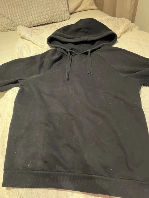 Svart hoodie från Gina Tricot - En svart hoodie från Gina Tricot med klassisk huva och dragsnören. Hoodien har långa ärmar och ribbade muddar vid ärmslut och nederkant. Perfekt för en avslappnad och stilren look.